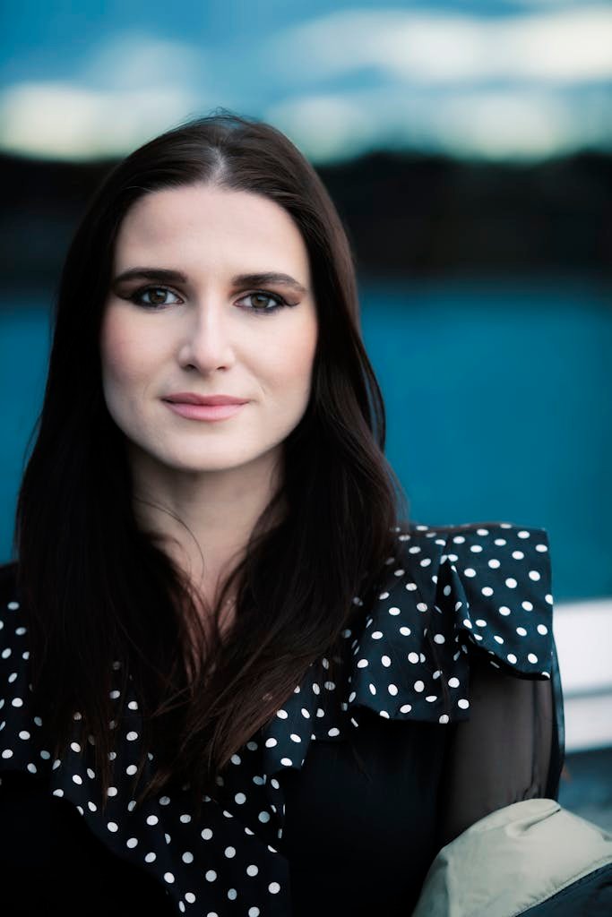 Portrait of a smiling woman with long dark hair in a polka dot blouse against a blurred background.