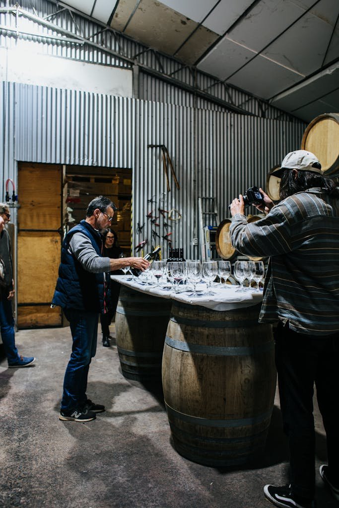 Full body of focused male pouring wine sample into wineglass at table with glassware while standing in winery with unrecognizable photographer